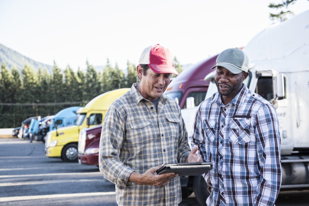 A caucasian man and a black man truck driving team together in a truck stop parking lot.